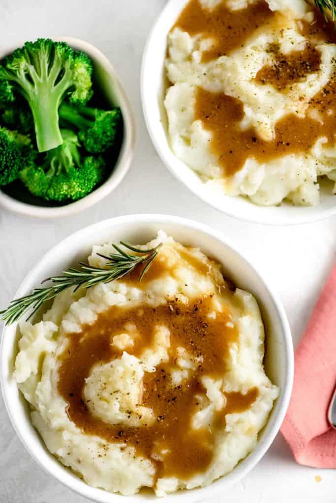 homemade mashed potatoes topped with ham gravy and rosemary beside a small bowl of broccoli