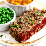 Thick cut of meatloaf with a rich ketchup glaze on a white plate beside a bowl of macaroni and cheese and green peas.