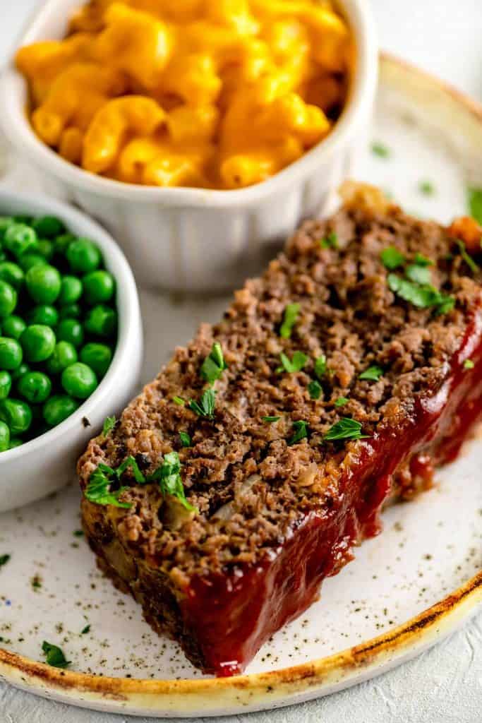 Thick cut of meatloaf with a rich ketchup glaze on a white plate beside a bowl of macaroni and cheese and green peas.
