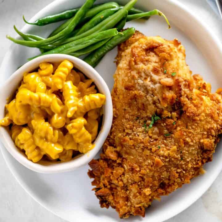 White plate with a giant crispy chicken breast next to macaroni and cheese and green beans.