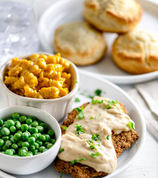 White plate with chicken fried steak covered in thick white pepper gravy beside peas, mac and cheese and biscuits