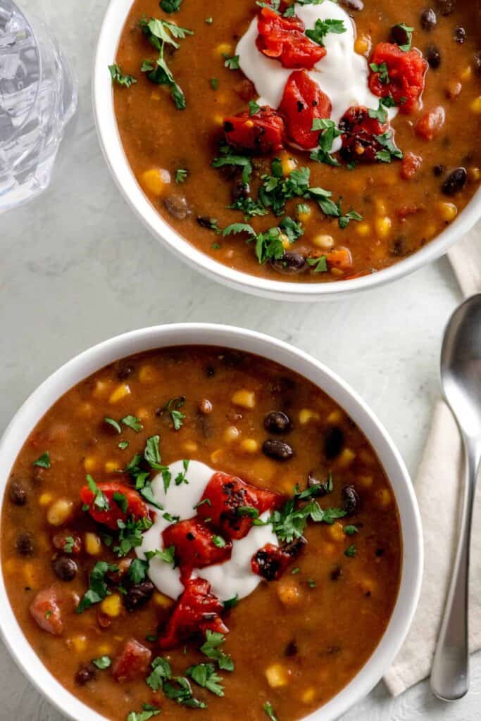 Two white bowls of thick bean soup topped with vegan yogurt, canned corn and canned tomatoes.