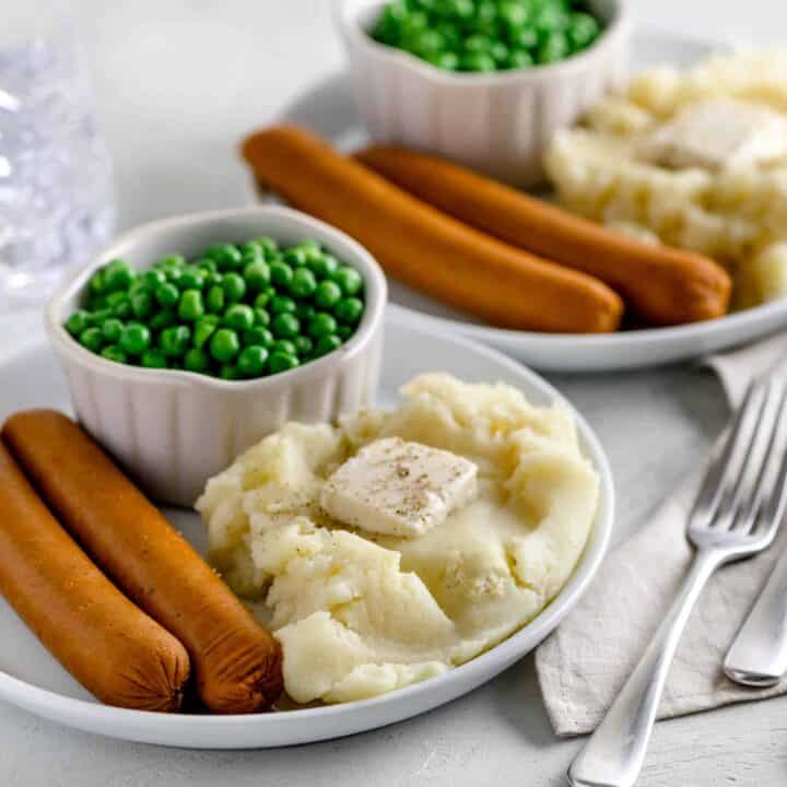 White plate with two vegan sausages, mashed potatoes topped with vegan butter and black pepper and a bowl of peas